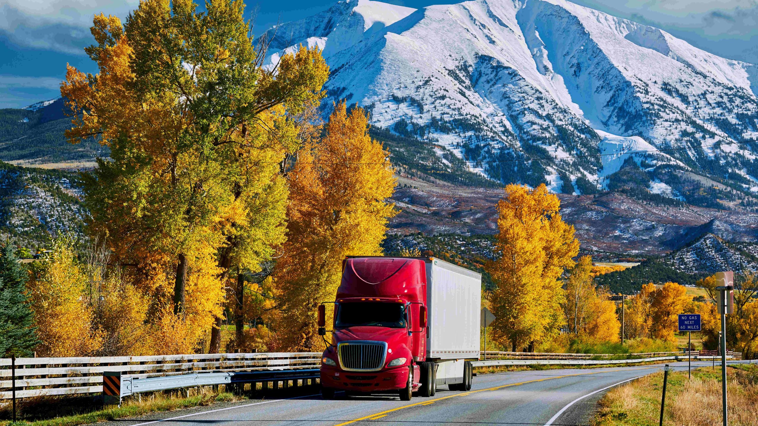 Red semi-truck for Managed Transportation services cruising through autumn foliage with snow-capped mountains in the background.