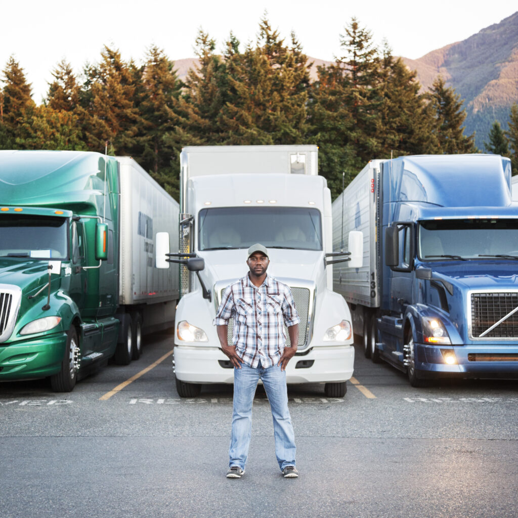 Less-than-Truckload driver standing confidently in front of three parked trucks in a scenic mountain setting.