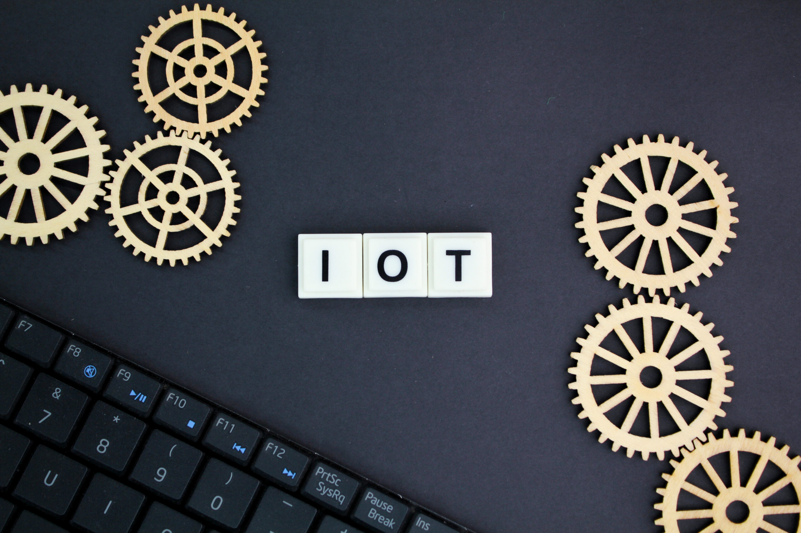 Keyboard with wooden gear wheels and the letters IOT spelled out in white blocks, representing Internet of Things in technology.