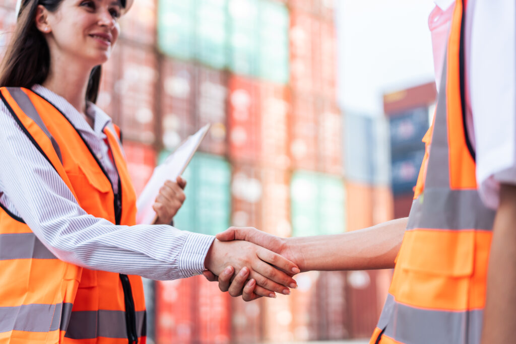 International business handshake between a businesswoman and a worker at a shipping yard, symbolizing global logistics and teamwork.