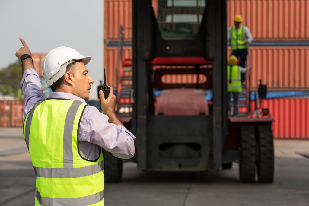 International engineer or foreman using walkie-talkie to communicate at a shipping yard, coordinating the movement of cargo containers.