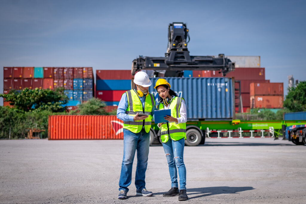 Two freight brokerage professionals in safety gear reviewing logistics plans at a shipping container yard.