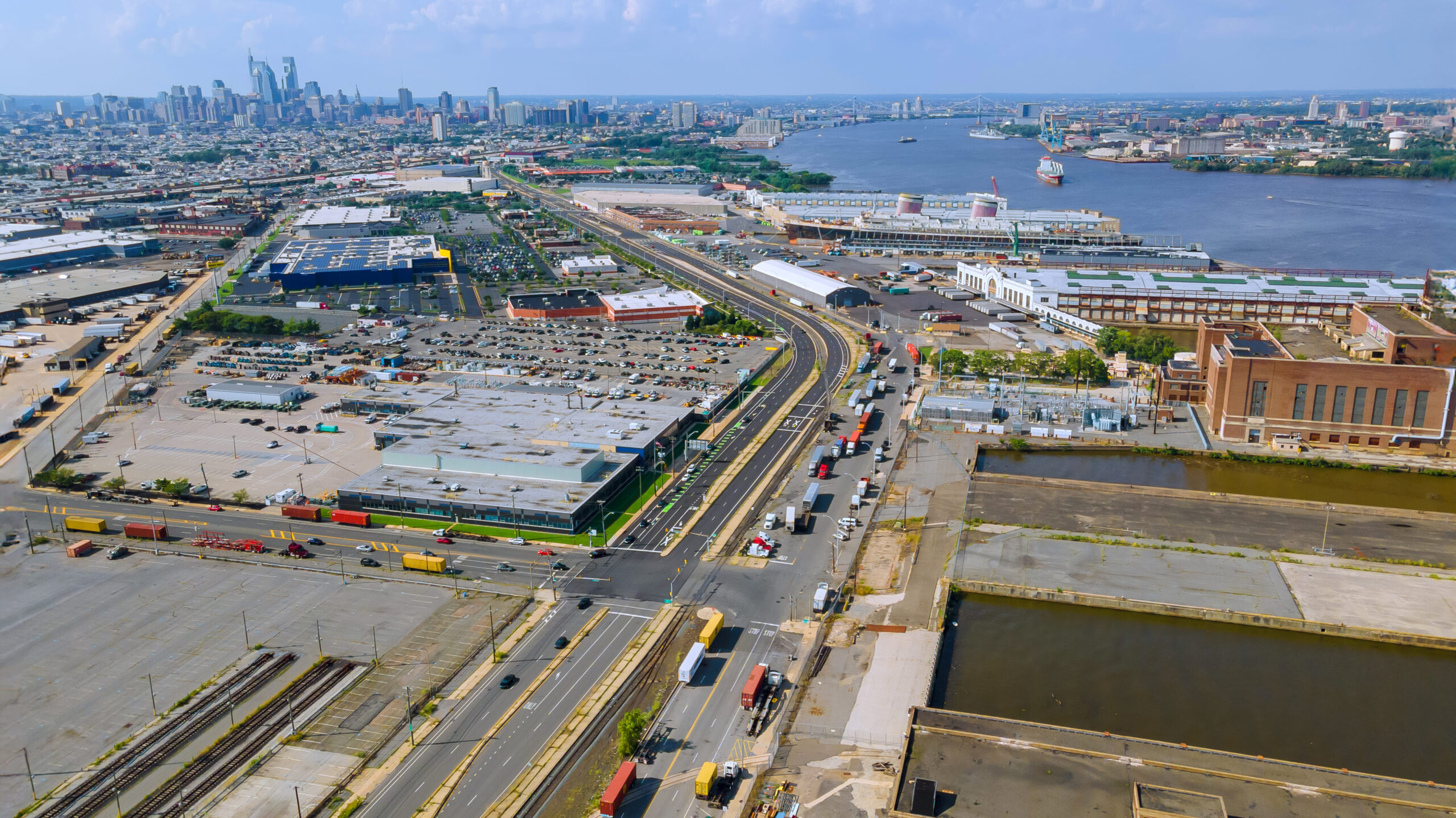 Aerial view of the Port of New York-New Jersey with cityscape in the background, showing shipping containers, trucks, warehouses, and waterfront infrastructure.