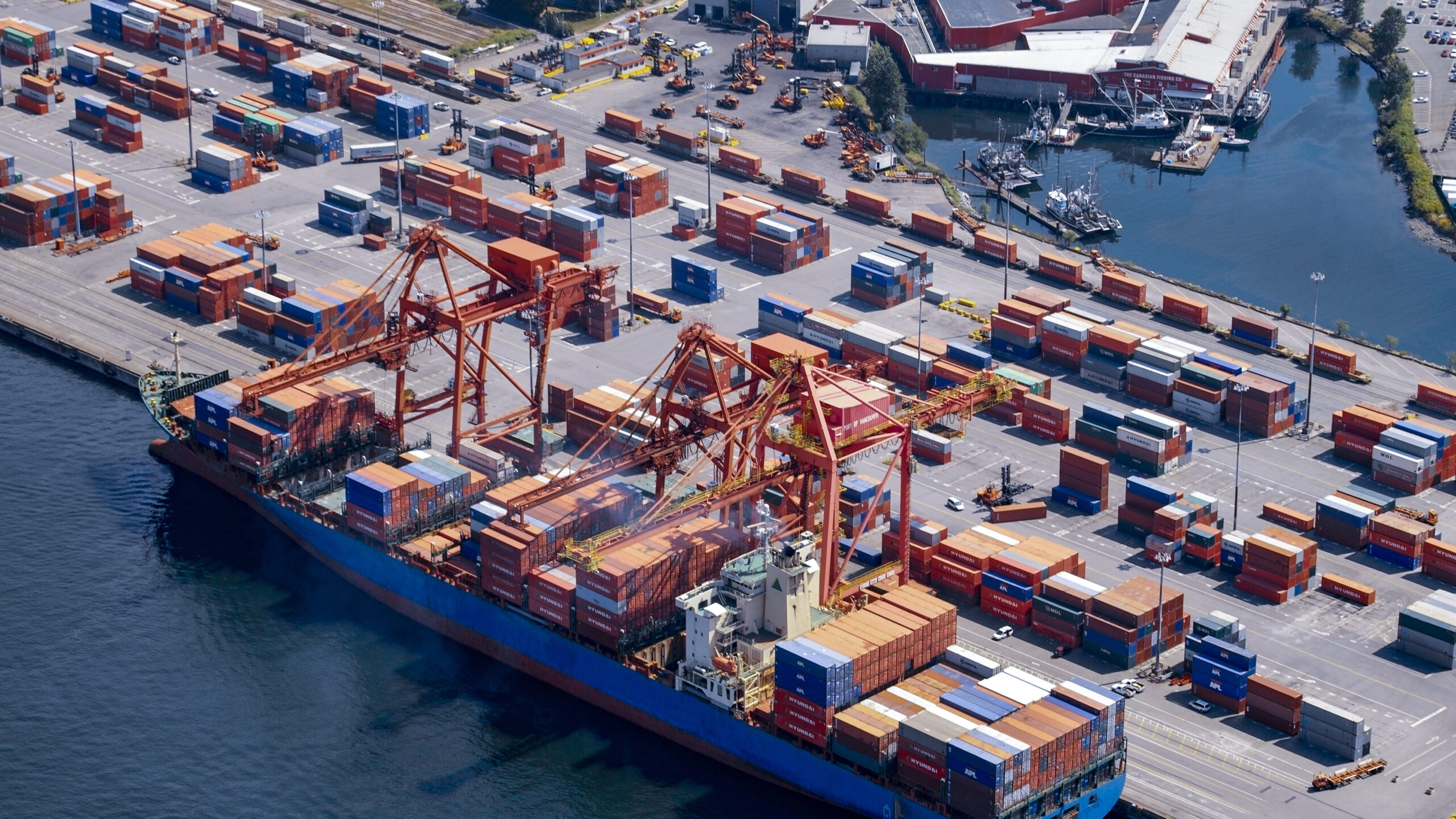 An aerial view of a large cargo ship docked at a busy container port, with numerous shipping containers and cranes, located at a coastal port facility.